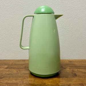 May include: A light green vintage thermos with a matching lid and handle. The thermos has a rounded base and a tall, slender body. The handle is a curved, rectangular shape. The lid is a dome shape. The thermos is made of plastic.