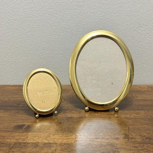 May include: Two gold-colored oval picture frames. The frames have a shiny finish and are supported by small, round feet. The smaller frame is on the left, and the larger frame is on the right. The frames are empty.