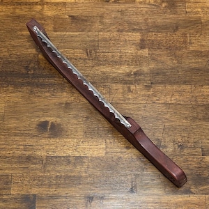 May include: A long, straight bread slicer featuring a dark brown wooden handle and a metallic, serrated blade attached with two screws. The tool is designed for cutting loaves of bread, with a long, narrow profile and a sharp, wavy edge.