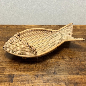 Puede incluir: Una cesta tejida en forma de pez de mimbre en tonos naturales. La cesta presenta un patrón de tejido detallado, con un cuerpo redondeado y una aleta caudal. El pez tiene un ojo decorativo y está diseñado para exhibición o almacenamiento. La cesta mide unos 51 cm de largo.