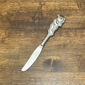 Peut inclure: Un couteau à beurre en métal argenté doté d'un manche décoratif représentant un ours grimpant sur un tronc d'arbre. Le manche présente une texture rappelant le bois, tandis que la lame est lisse, plate et se termine par une pointe arrondie.