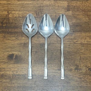 May include: Three silver-toned stainless steel spoons with a bamboo-inspired handle design. The leftmost spoon features a slotted bowl with four elongated openings, while the two spoons on the right have solid, smooth, reflective oval bowls.