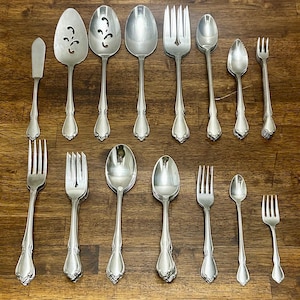 May include: A collection of vintage silver-plated flatware, including forks, spoons, and serving pieces. The set features ornate handles and a classic design. The pieces are arranged on a wooden surface, showcasing their reflective surfaces.