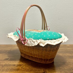 Puede incluir: Una cesta ovalada de mimbre trenzado con un asa alta arqueada. La cesta cuenta con una tapa acolchada cubierta de tela color turquesa brillante con un peque&ntilde;o patr&oacute;n floral blanco, adornada con encaje blanco y una cinta rosa fina.