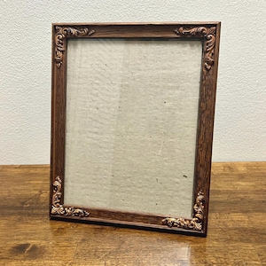 May include: A rectangular picture frame with a dark brown wood grain finish. The frame features ornate, raised gold-toned scrollwork detailing at each of the four corners. It contains a plain, light beige rectangular insert within the center.