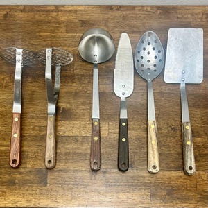 May include: A collection of vintage kitchen utensils, including a meat tenderizer, slotted spoon, ladle, cake server, and spatula. Each utensil has a stainless steel head and a wooden handle with brass rivets. The tools are arranged on a wooden surface.