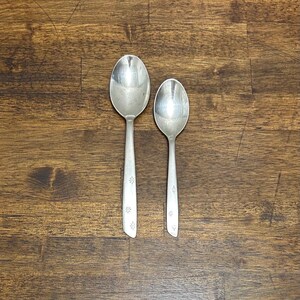 May include: Two silver spoons with a starburst design on the handles are displayed on a wooden surface. One spoon is larger than the other. The spoons are reflective and appear to be vintage.