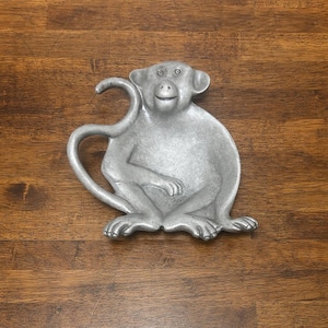 May include: A silver-colored monkey-shaped dish with a curled tail. The monkey is sitting with its arms resting on its legs. The dish is made of metal and has a detailed design. The dish is on a wooden surface.