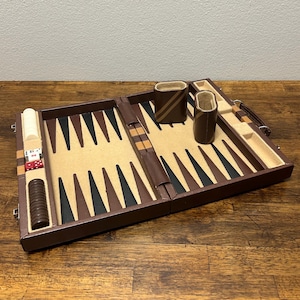 May include: A brown leather backgammon set, open and ready to play. The game board features alternating black, brown, and tan triangles. Two brown leather dice cups and game pieces are included. The set is on a wooden surface.