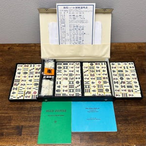 May include: A vintage Mahjong set with ivory-colored tiles, displayed in a black case. The set includes a variety of tiles with symbols and characters, along with instruction manuals and a paper with Japanese text. The case is open, revealing the organized arrangement of the tiles.