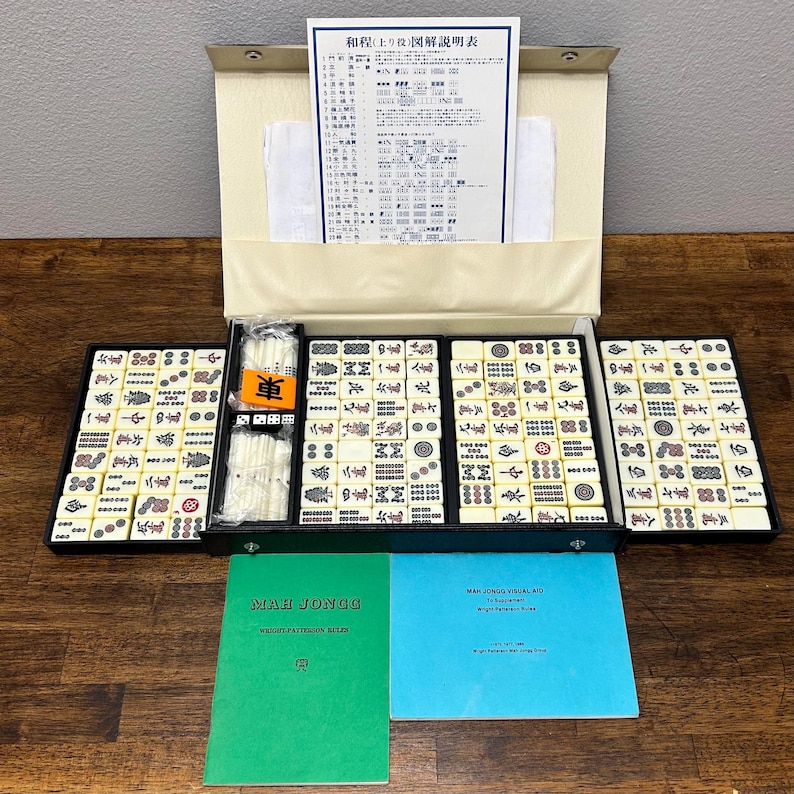 May include: A vintage Mahjong set with ivory-colored tiles, displayed in a black case. The set includes a variety of tiles with symbols and characters, along with instruction manuals and a paper with Japanese text. The case is open, revealing the organized arrangement of the tiles.