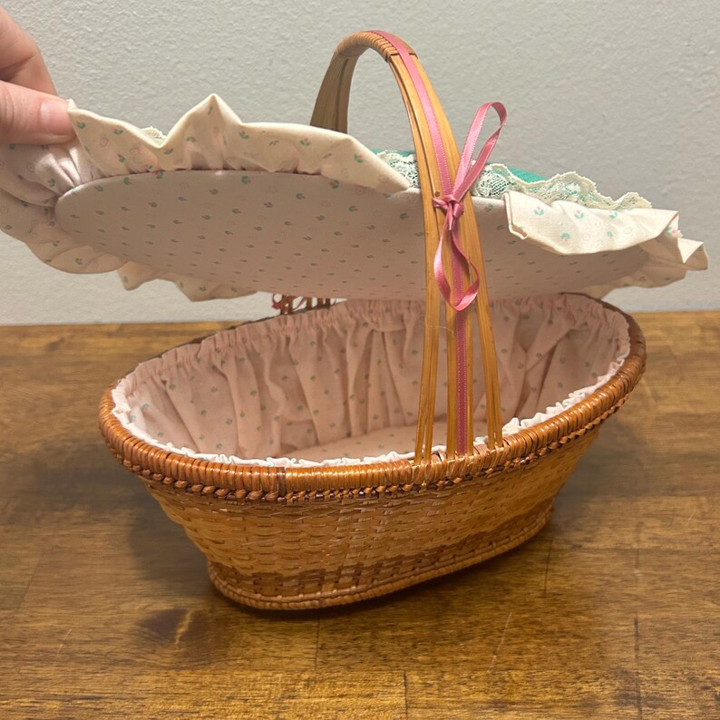 Puede incluir: Una cesta ovalada de mimbre trenzado con tapa abatible que presenta un forro de tela color crema con un peque&ntilde;o estampado floral verde. La tapa est&aacute; adornada con un borde de tela con volantes y encaje, y el asa tiene una fina cinta rosa.