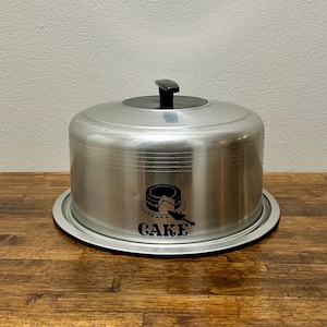 Könnte beinhalten: Ein Vintage-Kuchenbehälter aus Aluminium mit schwarzem Griff und Boden. Der Behälter hat eine schwarze Grafik eines Kuchens und das Wort "CAKE". Der Kuchenbehälter steht auf einer Holzoberfläche.