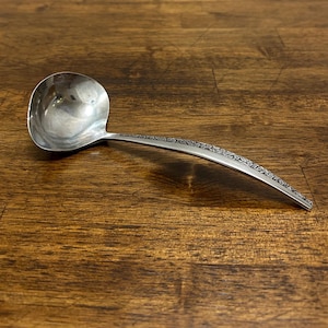 Puede incluir: Un cucharón plateado con un cuenco redondeado y un mango largo y curvado. El mango presenta un diseño floral decorativo. El cucharón está hecho de un metal brillante y reflectante y está sobre una superficie de madera oscura.