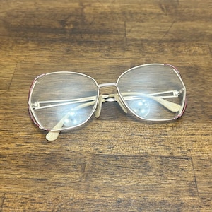 Könnte beinhalten: Brille mit silbernem Rahmen und klaren Gläsern. Die Fassungen haben eine abgerundete, übergroße Form mit dünnem Silbermetall und einem Hauch von Burgunder an den oberen Ecken. Die Bügel sind hellbeige. Die Brille liegt auf einer Holzoberfläche.