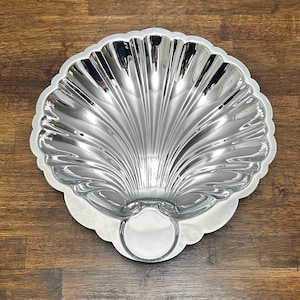 May include: A shiny, silver-colored seashell-shaped baking pan. The pan has a scalloped edge and a circular indentation at the base. The interior features a ribbed design, adding texture to the surface. This pan is designed for baking cakes or desserts.