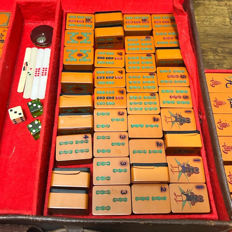 May include: A vintage Mahjong set featuring rectangular orange tiles with engraved green, blue, and black symbols, including bird illustrations and numbers. The set includes white counting sticks, green dice, and a dark circular token in a red-lined case.