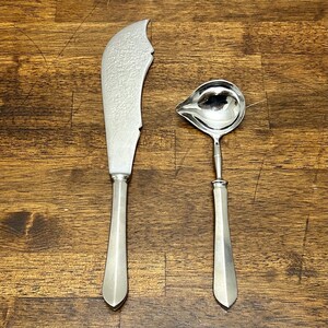 Puede incluir: Un juego de cuchillo de mantequilla y cucharón para salsa. El cuchillo de mantequilla tiene una hoja texturizada y un mango largo plateado. El cucharón para salsa tiene un cuenco redondeado y un mango largo plateado. Ambos utensilios son de metal brillante, sobre madera.