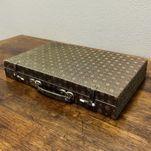 May include: A rectangular, closed case with a patterned fabric exterior. The fabric is a dark green with a repeating pink and gold design. The case has a silver handle and clasps.