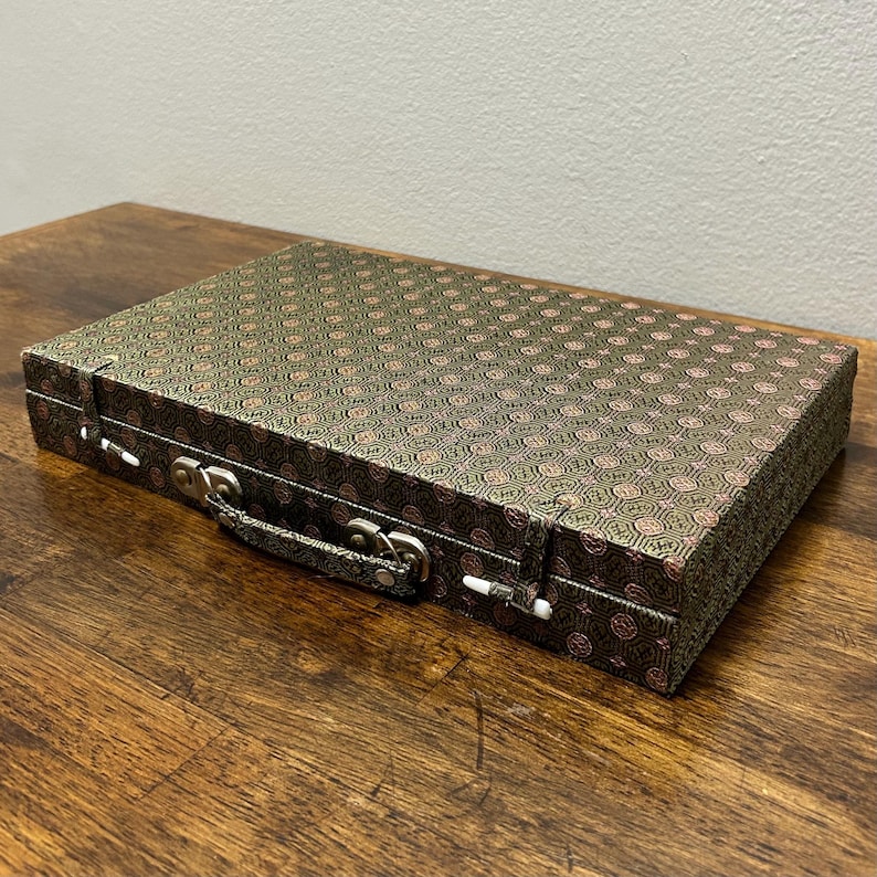 May include: A rectangular, closed case with a patterned fabric exterior. The fabric is a dark green with a repeating pink and gold design. The case has a silver handle and clasps.