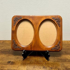 May include: A rectangular wooden double picture frame with a medium brown finish and visible wood grain. The frame features two oval-shaped openings and carved floral accents in each of the four corners. It is supported by a black stand.