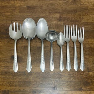 May include: A selection of vintage silverware, including serving spoons, forks, and a ladle, displayed on a wooden surface. The silverware has a shiny, silver finish and decorative detailing on the handles. The set includes a serving fork, spoons, and forks.