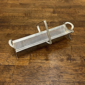 Puede incluir: Un rizador de mantequilla plateado con un diseño en forma de peine. El rizador tiene un asa y dos extremos curvos. Está diseñado para crear rizos de mantequilla decorativos. El rizador es de metal y está sobre una superficie de madera.