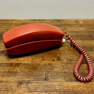 May include: A vintage red telephone with a coiled cord. The phone has a rectangular shape with rounded edges and a glossy finish. The cord is a matching red color and is coiled. The phone is sitting on a wooden surface.
