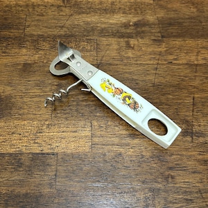 May include: A vintage combination bottle opener and corkscrew. The opener has a white handle with a decorative floral design in yellow, orange, and green. The metal components are silver-toned, and the tool is designed for opening bottles.