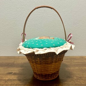 Puede incluir: Una cesta de mimbre marr&oacute;n tejida con una tapa de coj&iacute;n para alfileres de tela color turquesa con un peque&ntilde;o estampado floral. La tapa est&aacute; adornada con encaje blanco y un borde de tela con volantes, acentuada por dos lazos rosas y un asa fina.