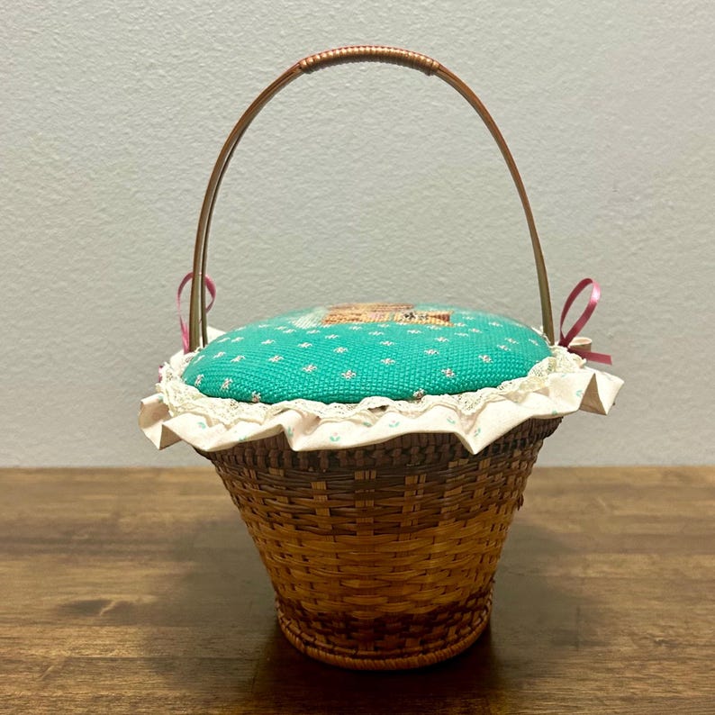 Puede incluir: Una cesta de mimbre marr&oacute;n tejida con una tapa de coj&iacute;n para alfileres de tela color turquesa con un peque&ntilde;o estampado floral. La tapa est&aacute; adornada con encaje blanco y un borde de tela con volantes, acentuada por dos lazos rosas y un asa fina.