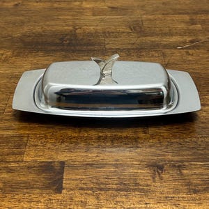 May include: A rectangular silver-toned metal butter dish with a polished, reflective finish. The dish features a matching lid with a V-shaped handle centered on top. The base has wide, flat edges extending from the central covered compartment.
