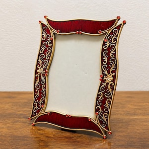 Puede incluir: Un marco de fotos decorativo con un diseño rojo y dorado. El marco tiene una abertura rectangular y está adornado con joyas rojas y adornos dorados. El marco es de metal y tiene una forma curva.