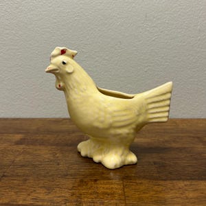 Könnte beinhalten: Ein hellgelber Keramik-Hühner-Pflanztopf. Der Pflanztopf hat einen roten Kamm und Kehllappen. Das Huhn hat ein detailliertes Design und steht auf einem strukturierten Sockel. Geeignet für kleine Pflanzen oder Blumen.
