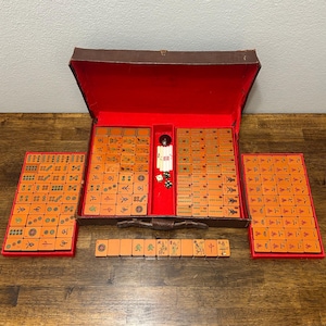 May include: A vintage Mahjong set featuring orange tiles with black and green engraved symbols. The set includes a brown carrying case with a red interior, two side trays, a row of tiles, and small white and green dice stored in the center.