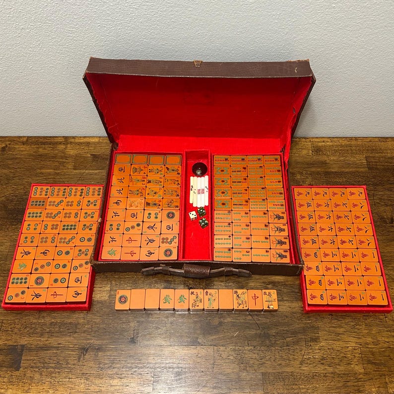 May include: A vintage Mahjong set featuring orange tiles with black and green engraved symbols. The set includes a brown carrying case with a red interior, two side trays, a row of tiles, and small white and green dice stored in the center.
