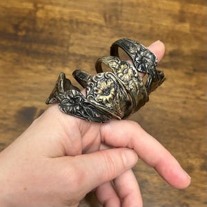 May include: A set of three silver-toned metal rings featuring an embossed floral pattern with dark oxidized detailing. The rings have a wide, curved band design with a textured finish, showcasing intricate petal and leaf motifs across the surface.