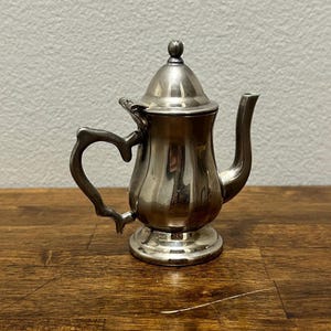 May include: A small, silver-colored teapot with a lid and a curved handle. The teapot has a spout and sits on a round base. The handle is heart-shaped. The teapot is on a wooden surface.