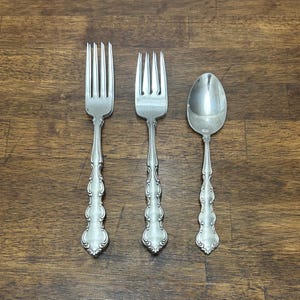 Può includere: Tre posate d'argento: due forchette e un cucchiaio. Le posate sono di colore argento con manici decorativi e ornati. Le forchette hanno quattro rebbi ciascuna. Il cucchiaio ha una coppa arrotondata. Le posate sono disposte su una superficie di legno.