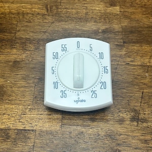 Puede incluir: Un temporizador de cocina cuadrado de color blanco con bordes redondeados y un dial giratorio central. La cara muestra marcas numéricas negras del 0 al 55 en incrementos de cinco minutos, con la marca UpJate impresa en negro abajo al centro.