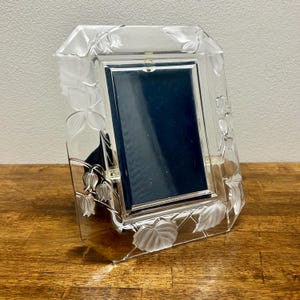 May include: A rectangular clear glass picture frame featuring frosted floral and leaf relief carvings along the border. The frame has a central vertical opening with a dark blue backing and a metallic silver-toned inner trim surrounding the display area.