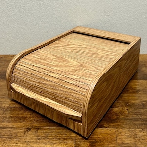 Peut inclure: Une boîte à pain en bois avec un couvercle roulant. La boîte est en bois brun clair avec un motif de grain. La boîte est rectangulaire avec des bords arrondis et repose sur une surface en bois.
