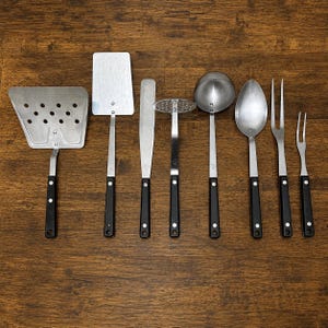May include: A set of eight stainless steel kitchen utensils with black handles and silver rivets, arranged in a row. The collection includes a slotted turner, a solid turner, a spatula, a potato masher, a ladle, a spoon, and two meat forks.
