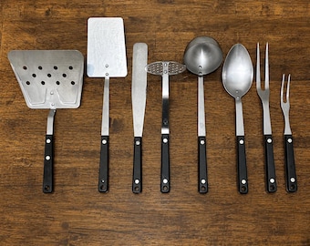 Espátula, machacador de patatas, cucharón o tenedor para trinchar de la marca Ekco Forge, con mango de material compuesto negro y punta de flecha de sílex. Fabricada en acero inoxidable. Se vende por unidad.