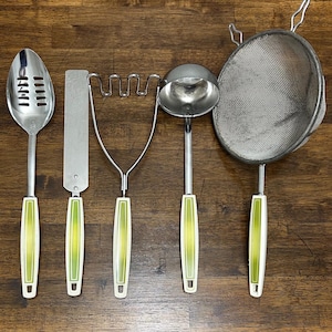 Puede incluir: Un juego de seis utensilios de cocina vintage con cabezales de acero inoxidable y mangos blancos y verdes. Incluye una cuchara ranurada, espátula, pasapurés, cucharón y colador de malla.