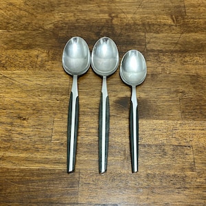 Puede incluir: Tres cucharas plateadas con mangos negros y blancos dispuestas sobre una superficie de madera. Las cucharas tienen un diseño clásico con cuencos ovalados y mangos delgados. Los mangos son negros con una raya blanca.