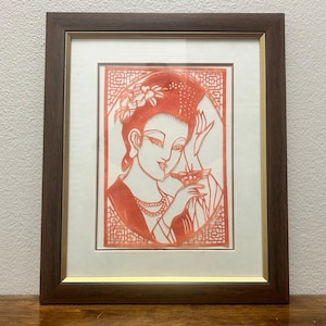 May include: A framed paper cut art piece featuring a coral-colored silhouette of a person with an elaborate hairstyle, holding a small cup. The artwork is set against a white mat with a gold inner border, enclosed in a dark brown wooden frame.
