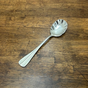 May include: A silver serving spoon with a decorative, scalloped bowl and a long, slender handle. The spoon is made of a shiny, reflective metal and rests on a dark wooden surface. The spoon is designed for serving food.