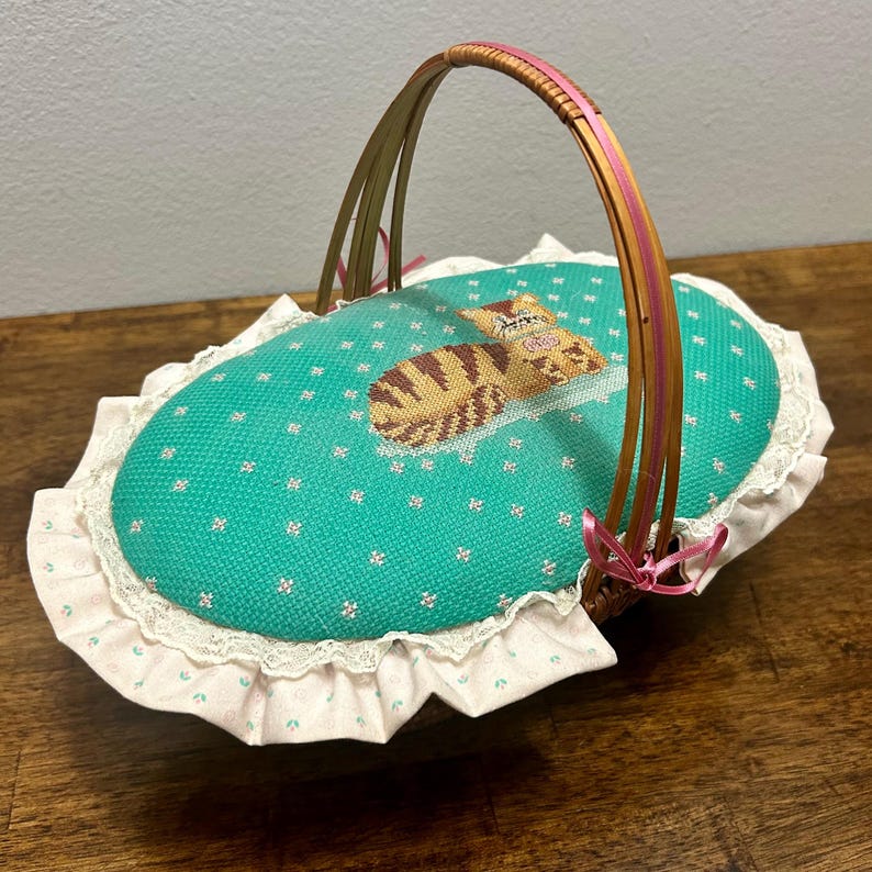Puede incluir: Una cesta de costura ovalada con una tapa de tela turquesa que presenta un gato atigrado marr&oacute;n bordado en punto de cruz. La tapa est&aacute; bordeada con encaje blanco y un volante floral rosa claro. Un asa de madera arqueada tiene una cinta rosa.