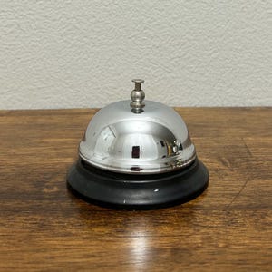 May include: A shiny, chrome-plated bell with a black base sits on a wooden surface. The bell has a rounded top and a small, decorative handle. The bell's surface reflects the surrounding environment, creating a mirrored effect.